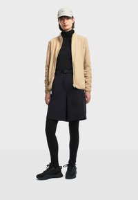 Veste zippée en polaire beige, haut à manches longues noir, short noir avec ceinture, leggings noirs et baskets noires. Casquette de couleur claire sur la tête.