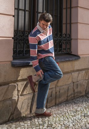 Uomo con maglia da rugby a righe e jeans che sistema la sua scarpa marrone mentre si trova in una strada lastricata vicino a un muro di pietra.