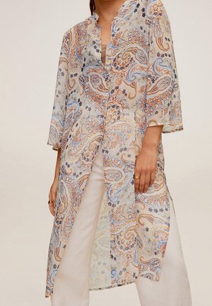Kvinde iført en lang, transparent bluse med paisley-mønster i pastelfarver over hvide bukser, med ærmer, der går lige under albuerne.