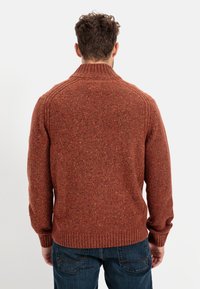 Ein rostfarbener gestrickter Pullover mit hohem Kragen, gerippten Bündchen und Saum. Strukturierter Stoff mit Farbklecksen überall.