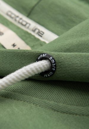 Vihreä puuvillakangas, jossa on teksturoitu pinta, sisältää valkoisen narun, joka on kiinnitetty mustaan muovilenkkiin, jossa lukee "wear ombre."