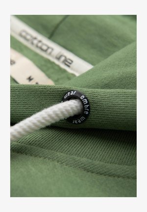 Grüner Baumwollstoff mit einer strukturierten Oberfläche, versehen mit einer weißen Kordel, die durch einen schwarzen Kunststofföse gesichert ist, die mit "wear ombre" beschriftet ist.