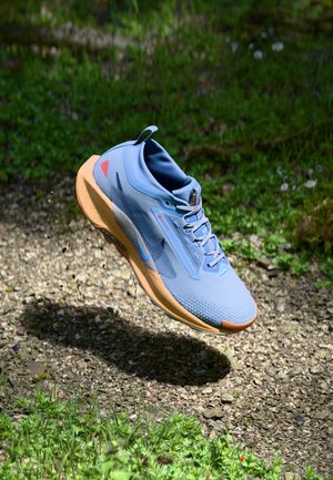Blauwe sportschoen met een gemetalliseerde bovenkant van mesh, rubberen zool en oranje accenten, weergegeven in de lucht tegen een natuurlijke achtergrond.