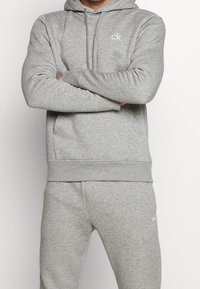 Felpa grigia con cappuccio e pantaloni abbinati, realizzati in un tessuto morbido. Presenta un cappuccio con cordino e tasche frontali; piccolo logo "ck" sul petto.