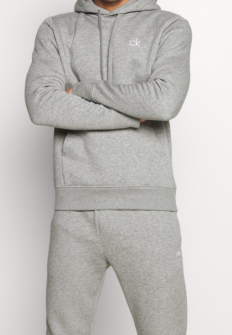 Felpa grigia con cappuccio e pantaloni abbinati, realizzati in un tessuto morbido. Presenta un cappuccio con cordino e tasche frontali; piccolo logo "ck" sul petto.