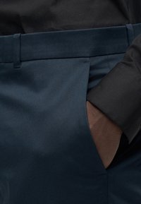 Μπλε μπλε παντελόνι από λείο ύφασμα με επίπεδη μπροστινή όψη, που διαθέτει πλαϊνή τσέπη και μερικώς ορατό μανίκι μαύρου πουκαμίσου.