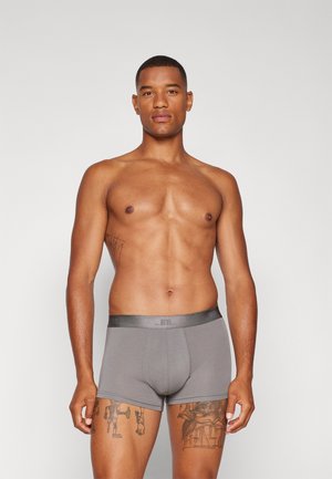 Grå boxershorts med en åtsittande design, slät textur och en kontrasterande midjeband. Har tatueringar synliga på låren.