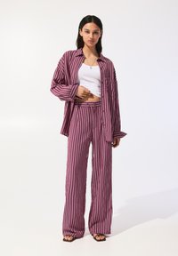 Pruhovaný dvoudílný outfit v tmavě vínové a bílé barvě. Volná dlouhá košile s dlouhým rukávem a široké kalhoty s hladkou texturou. Černé sandály.
