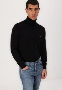 Pulover negru cu guler înalt și mâneci lungi, având un mic logo galben pe pieptul stâng, asortat cu blugi albaștri.