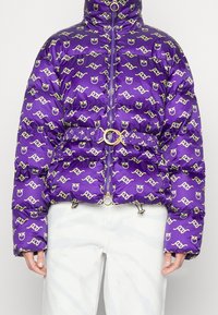 Paarse donsjas met overal een beige logopatroon, ritssluiting aan de voorkant, ceintuur met gouden ringgesp in de taille, gedragen met witte broek.