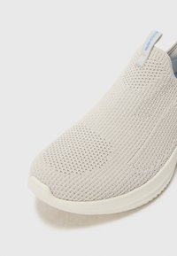 Chaussure slip-on gris clair avec une tige en tricot respirant, un motif texturé et une semelle blanche. Présente une petite étiquette d'accents bleus sur le talon.