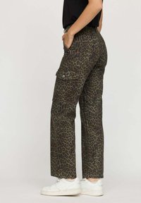 Pantalons cargo vert olive avec imprimé léopard noir. Comprend des poches latérales, une coupe droite et un tissu durable. Des baskets blanches sont portées.