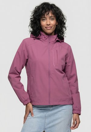 Frau mit lockigem Haar, die eine lila Kapuzenjacke und einen hellblauen Jeansrock trägt, steht mit einer Hand in der Hüfte.