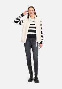 Weste aus warmem, weißem Fleece über einem schwarz-weiß gestreiften Pullover, kombiniert mit dunklen Jeans und schwarzen Stiefeletten. Einfaches Design, keine auffälligen Muster.