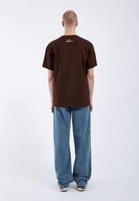 Camiseta de algodón marrón con mangas cortas, que presenta un logotipo blanco en la parte posterior, combinada con unos vaqueros de denim azules de corte holgado.