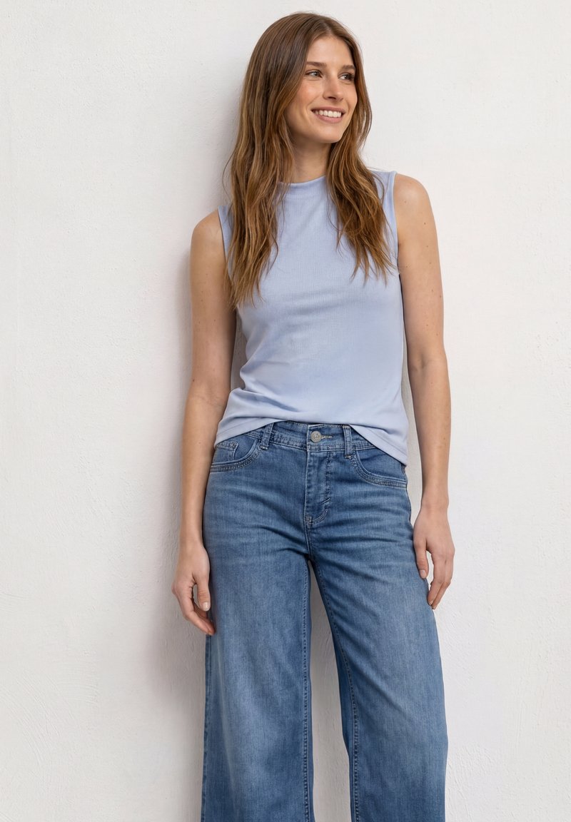 Jeune femme souriante, portant un haut sans manches bleu clair et un jean bleu large taille haute, debout contre un mur blanc uni.
