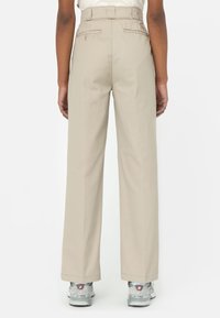 Zadní pohled na osobu v béžových kalhotách s vysokým pasem a zadními kapsami, která má na sobě šedé tenisky New Balance na bílém pozadí.