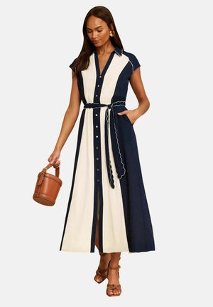 Donna che indossa un vestito midi blu navy e bianco con bottoni frontali e maniche a cappuccio, che tiene una borsa a secchiello marrone e indossa sandali marroni con tacco.