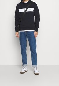 Osoba w czarno-białym hoodie, niebieskich dżinsach z podwiniętymi nogawkami, białych sneakersach z czarnymi paskami i czarnej torbie na ramię na białym tle.