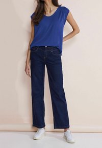 Blue short-sleeve top with a round neckline paired with dark blue wide-leg jeans and white sneakers, set against a neutral background.