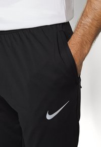Schwarze Sporthose mit elastischem Bund, seitlichen Taschen und einem silbernen Nike-Logo auf dem linken Oberschenkel. Glatter, leichter Stoff.
