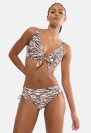 Γυναίκα που φοράει μπικίνι με ρίγες τίγρης, το πάνω μέρος έχει κόμπο μπροστά και το κάτω μέρος δένεται στο πλάι, στέκεται μπροστά σε απλό φόντο.