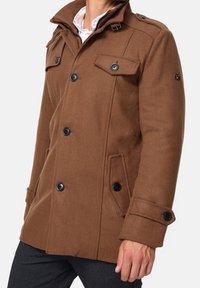 Bruine jas van een wollen mix met een dubbelbreasted ontwerp, zwarte knopen, schouderepauletten en zijzakken. Hoge kraag met verstelbare riem.