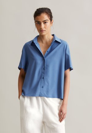 Blaues kurzärmliges Button-up-Hemd aus glattem Stoff mit klassischem Kragen und Vorderknöpfen; kombiniert mit weißen, locker sitzenden Hosen.