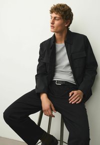 Jeune homme aux cheveux bouclés assis sur un tabouret, portant une veste noire, une chemise grise, un pantalon noir et des chaussures noires, regardant sur le côté contre un mur blanc.