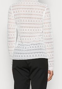 Vit spets långärmad skjorta med ett blommönster och en texturerad design, tillsammans med svarta byxor. Baksidan visar intrikat spetsarbete.