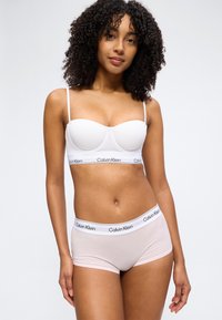 Femeie cu păr creț purtând sutien alb Calvin Klein și chiloți roz deschis Calvin Klein, în picioare pe un fundal simplu.