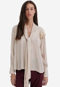 Lichtbeige blouse met een strikcol, lange mouwen, textuurstof en bloemendetails op de linkerschouder, gecombineerd met donker bordeauxrode broeken.