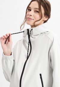 Leichte, cremefarbene Jacke mit schwarzem Reißverschluss und Kordelzug. Verfügt über elastische Bündchen und eine Reißverschlusstasche für zusätzlichen Komfort.