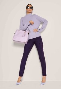 Frau in lilafarbenem Rollkragenpullover, dunkellila Hose, lilafarbenen High Heels, großer lilafarbener Handtasche und übergroßen Sonnenbrille, die selbstbewusst posiert.