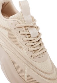 Beige Sneaker mit einem strukturierten Mesh-Obermaterial, glatten Gummi-Akzenten, flachen Schnürsenkeln und einem gepolsterten Zungenbereich für zusätzlichen Komfort.