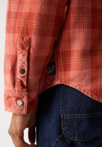 Camisa de cuadros roja y rosa con un puño abotonado, que presenta un detalle de botón redondo azul y blanco. La tela muestra un tejido texturizado y costuras.
