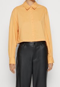 Chemise cropped à manches longues en tissu orange doux avec un devant boutonné et un col doux, associée à un pantalon en cuir noir taille haute.