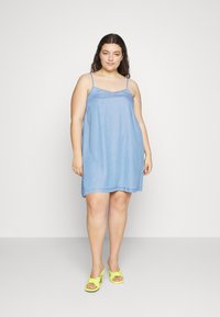 Vestido de ganga azul claro com tiras finas, em forma A, e uma textura suave; combinado com sapatos de salto amarelos brilhantes.