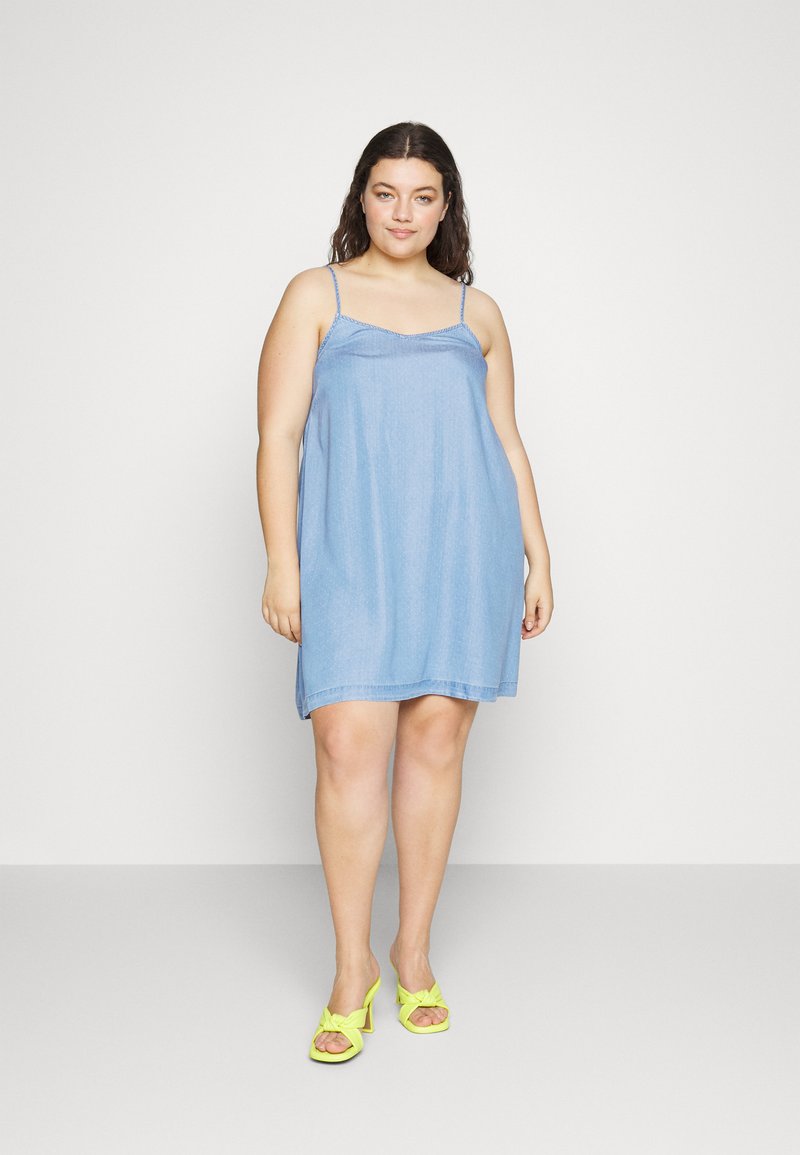 Vestido de ganga azul claro com tiras finas, em forma A, e uma textura suave; combinado com sapatos de salto amarelos brilhantes.