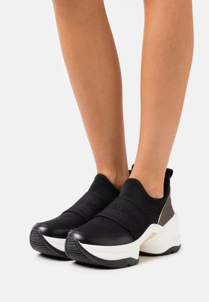 Persona que lleva zapatillas negras sin cordones con suelas blancas gruesas, talón texturizado y una amplia banda elástica cruzando el pie.