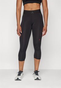 Svarte capri leggings med en glatt tekstur, featuring en elastisk midje og subtil branding i rødt på siden. Kombinert med hvite sportssko.