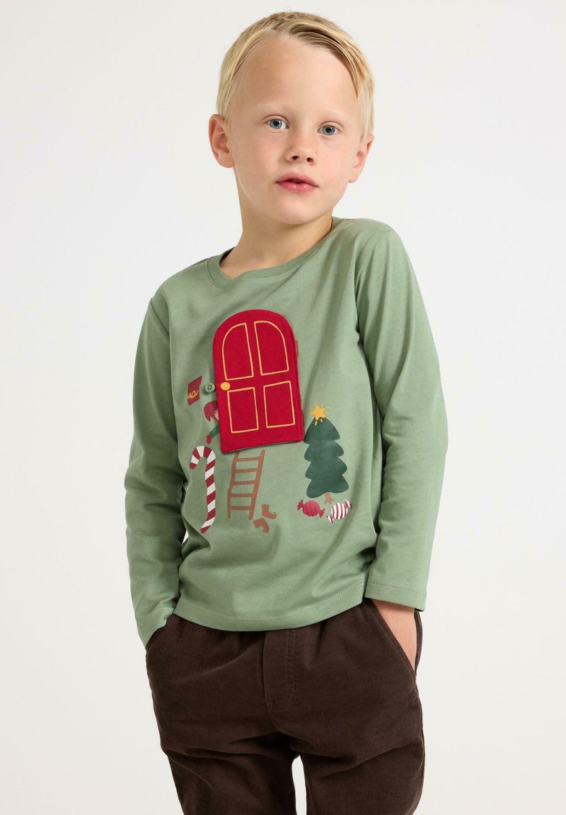 Grøn langærmet skjorte med en rød dør-appliqué, et juletræ, slikstokke og et stige-tryk. Parret med brune fløjlbukser.