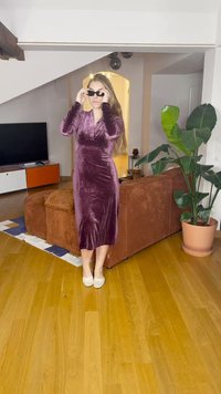 Bársonyos, mély lila ruha hosszú ujjakkal, amely öleli a sziluettet, fekete napszemüveggel párosítva. Fán burkolt padlón áll, közel egy növényhez.