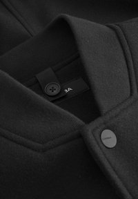 Črna volnena jakna s zloženim ovratnikom, ki se zapira na gumb in ima etiketo iz blaga. Gladka tekstura z minimalnimi detajli.