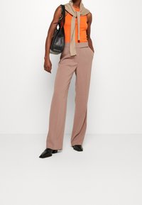 Beige vida byxor med texturerad tyg, i kombination med en orange topp. Outfiten har en beige tröja draperad över axlarna och en svart väska.