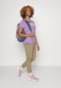 Camiseta morada con logotipo verde, pantalones beige y zapatillas rosa claro. La mochila tiene un patrón floral, tirantes ajustables y un bolsillo con cremallera.