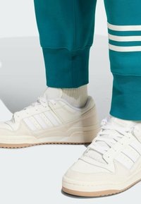 Vita sneakers i texturerat läder och mockadetaljer, med tre horisontella ränder. Kombinerat med teal joggers med vita ränder.
