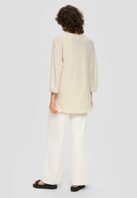 Béžový top s dlouhým rukávem a volným střihem v kombinaci s bílými kalhotami s širokými nohavicemi. Černé platformové sandály dokončují outfit.