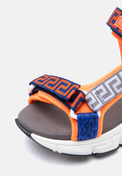 Πέδιλο ανοιχτού δακτύλου με πορτοκαλί ύφασμα, ρυθμιζόμενα λουριά με ναυτικό μπλε και λευκό σχέδιο, γκρι επενδυμένο πάτο και λευκή σόλα με το λογότυπο "Versace".