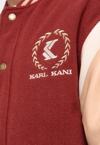 Bordowa kurtka varsity z wełny z beżowymi skórzanymi rękawami, z haftowanym logo i napisem. Przód zdobią złocone zatrzaski.